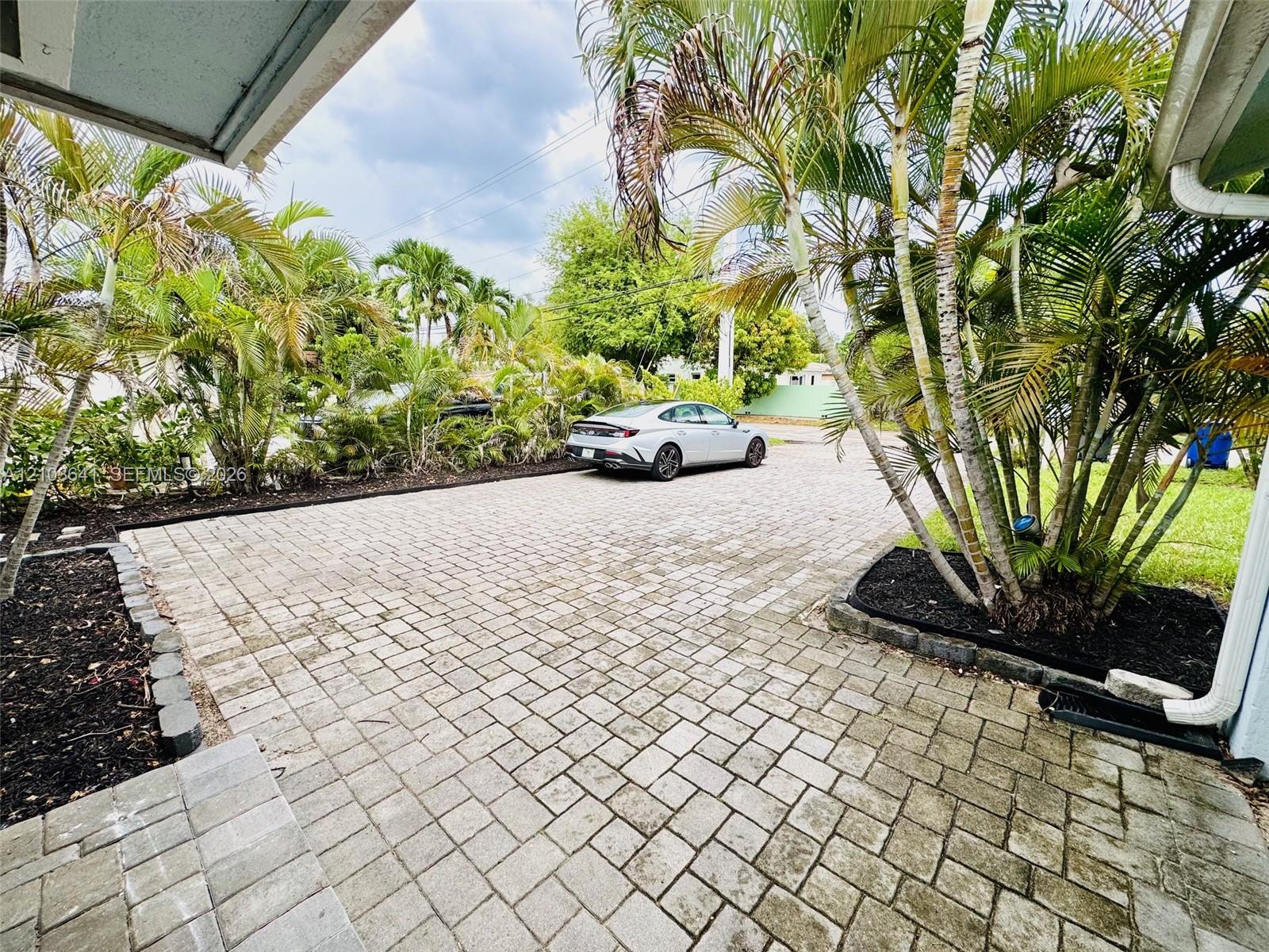 Paver driveway featuring stone-finished hardscaping, manicured garden beds with dark mulch, and established palm trees