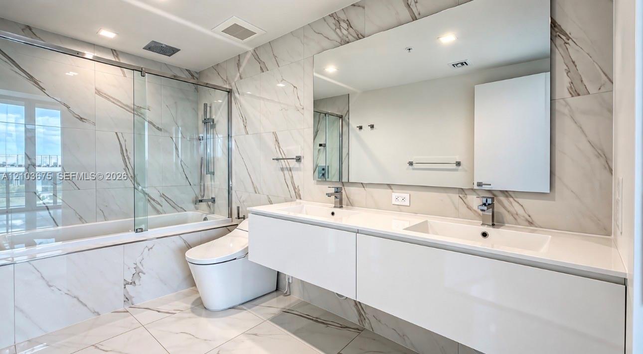 Floating dual-sink vanity with a solid surface countertop, large wall-mounted mirror, floor-to-ceiling stone-patterned tile, and a glass-enclosed shower and tub combination