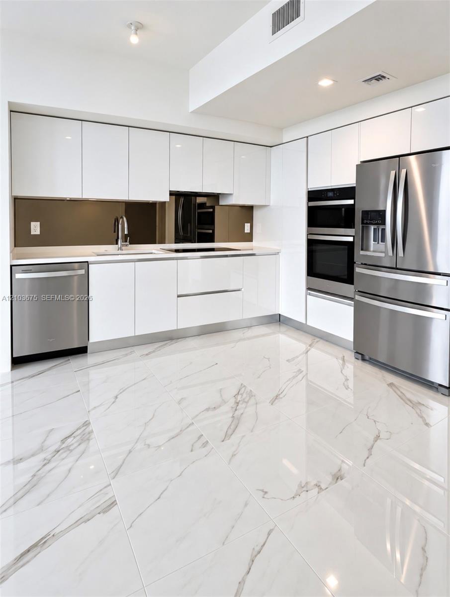 High-gloss flat-panel white cabinetry, stainless steel appliances, integrated double wall ovens, mirror-finish backsplash, and polished stone-finish tile flooring