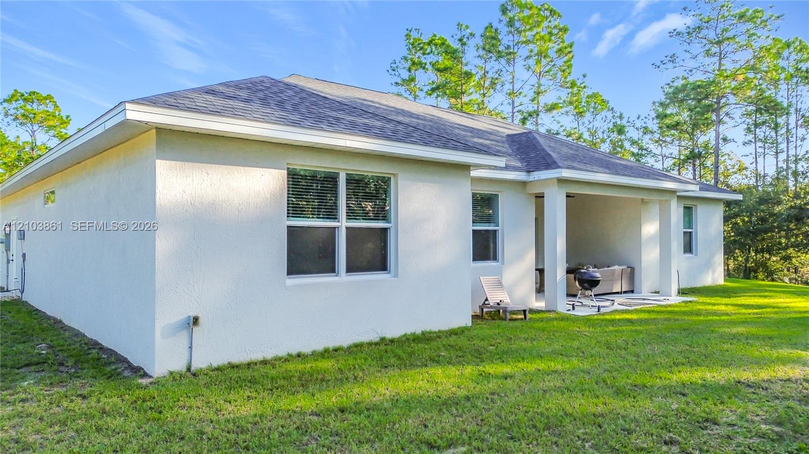 Stucco exterior featuring a covered patio, shingle roof, and expansive backyard with mature pine trees