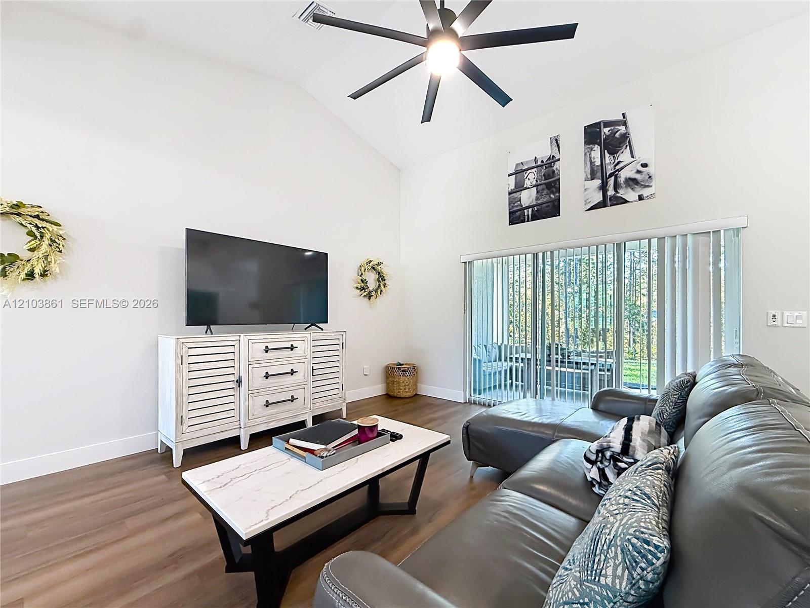 Vaulted ceiling featuring a central matte black ceiling fan, wood-finish flooring, and sliding glass patio doors with vertical blinds