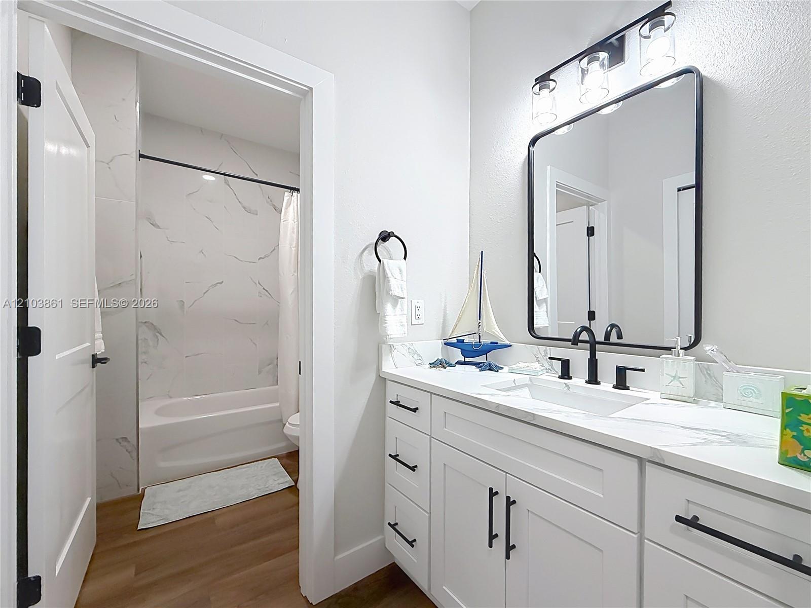 Bathroom featuring a white vanity with stone-style countertop, rectangular undermount sink, matte black hardware, framed vanity mirror, and tub-shower combination with floor-to-ceiling tile surround