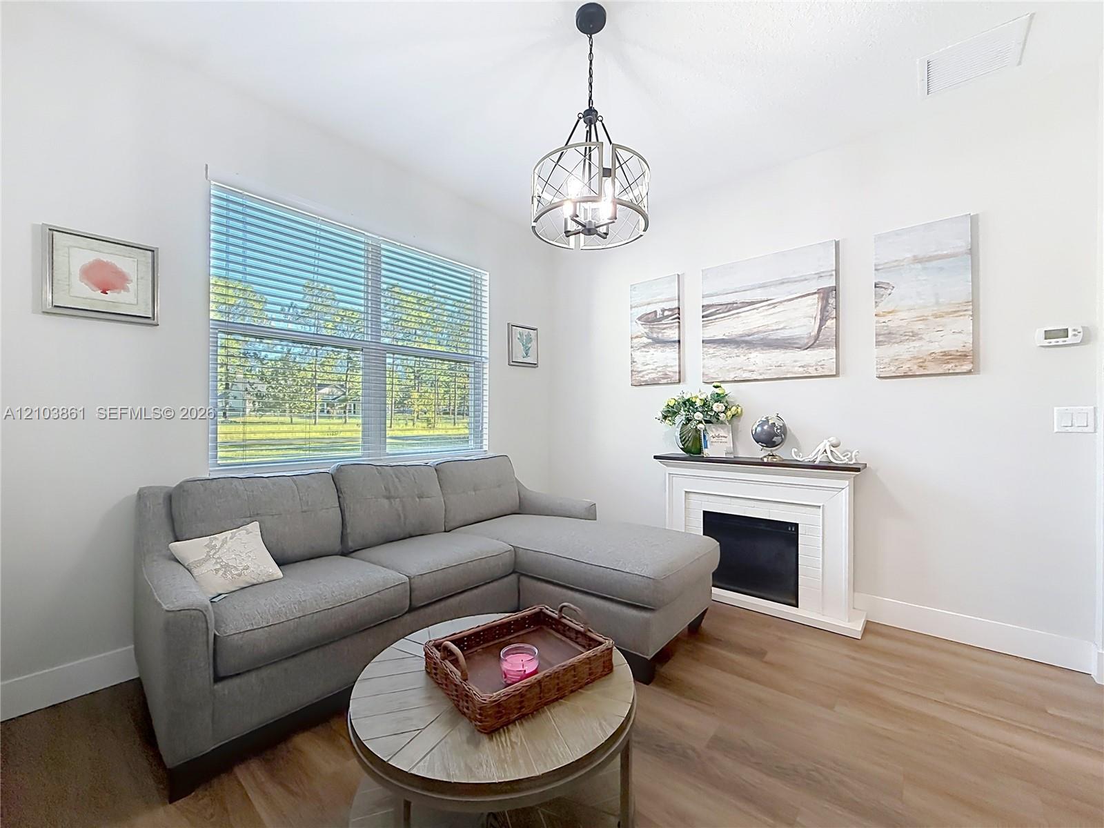 Wood-finish flooring, white fireplace surround with integrated mantel, large window with horizontal blinds, and a multi-light metal pendant fixture