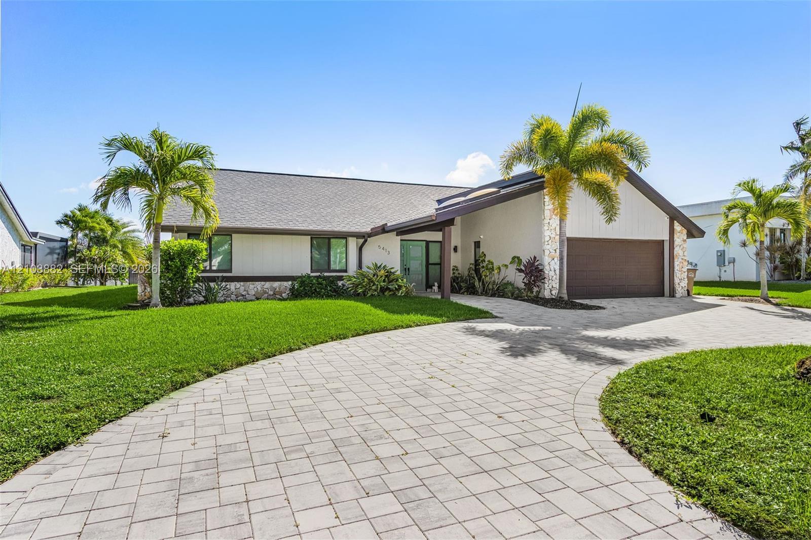 Single-story exterior featuring a stone-clad facade, paver driveway, gabled roofline, integrated garage, and tropical landscaping
