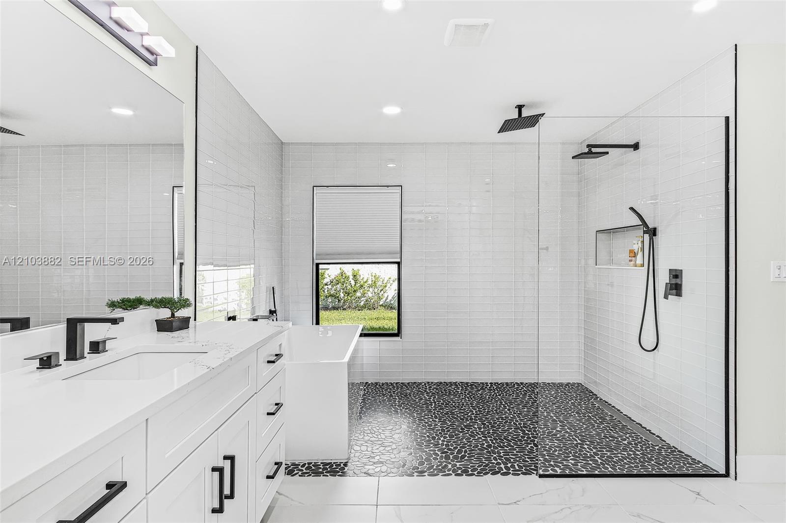 White vanity with stone countertop, freestanding bathtub, walk-in shower with river stone flooring, glass enclosure, and matte black fixtures