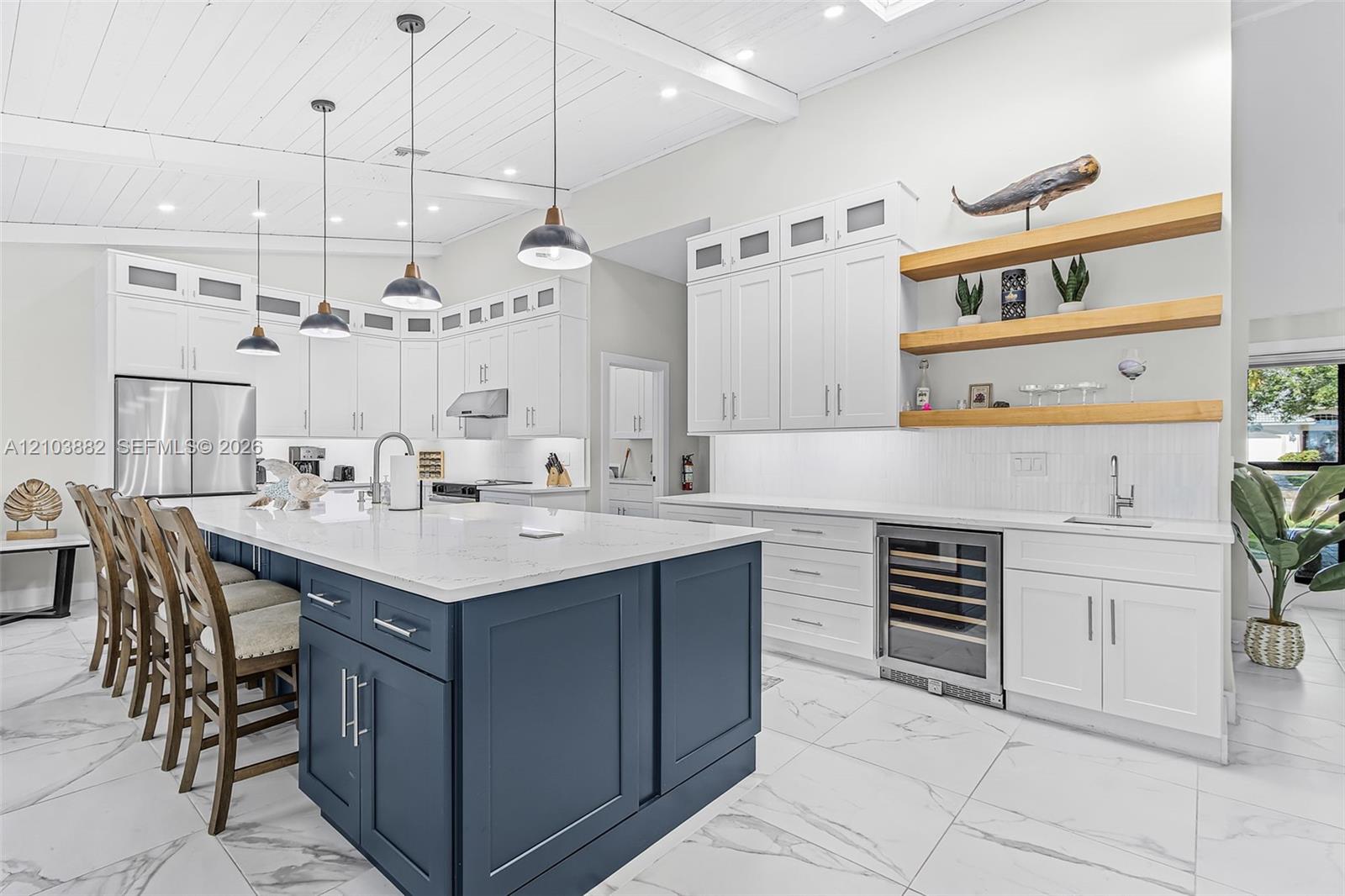 Kitchen featuring a large blue center island with stone-finish countertops, ceiling-height white cabinetry, built-in stainless steel wine refrigeration, natural wood-finish open shelving, and tongue-and-groove ceiling detail