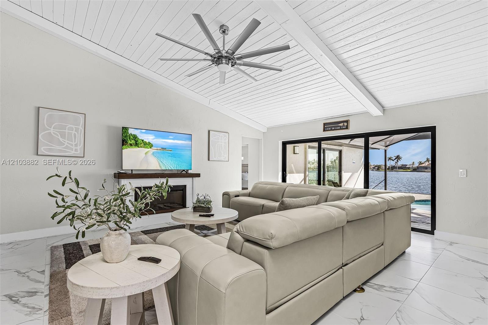 Vaulted tongue-and-groove ceiling, expansive multi-panel sliding glass doors, fireplace with a wood mantel, and large-format white stone-finish flooring