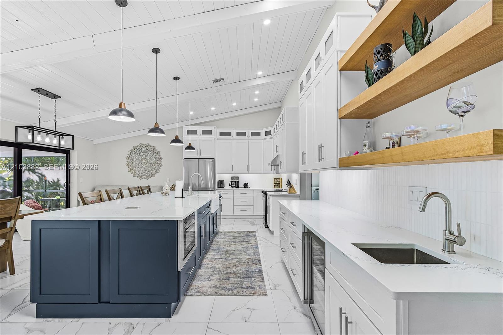 Kitchen featuring white shaker-style cabinetry, a large navy-toned center island with light-toned stone-finish countertops, exposed wood shelving, and a vaulted ceiling with exposed beams