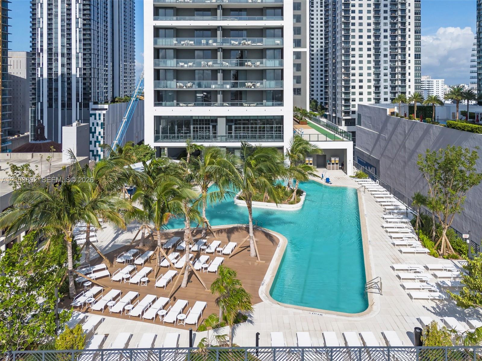 Elevated outdoor pool with a sun shelf, wood-finish pool decking, surrounding palm trees, and light-toned stone paved terrace