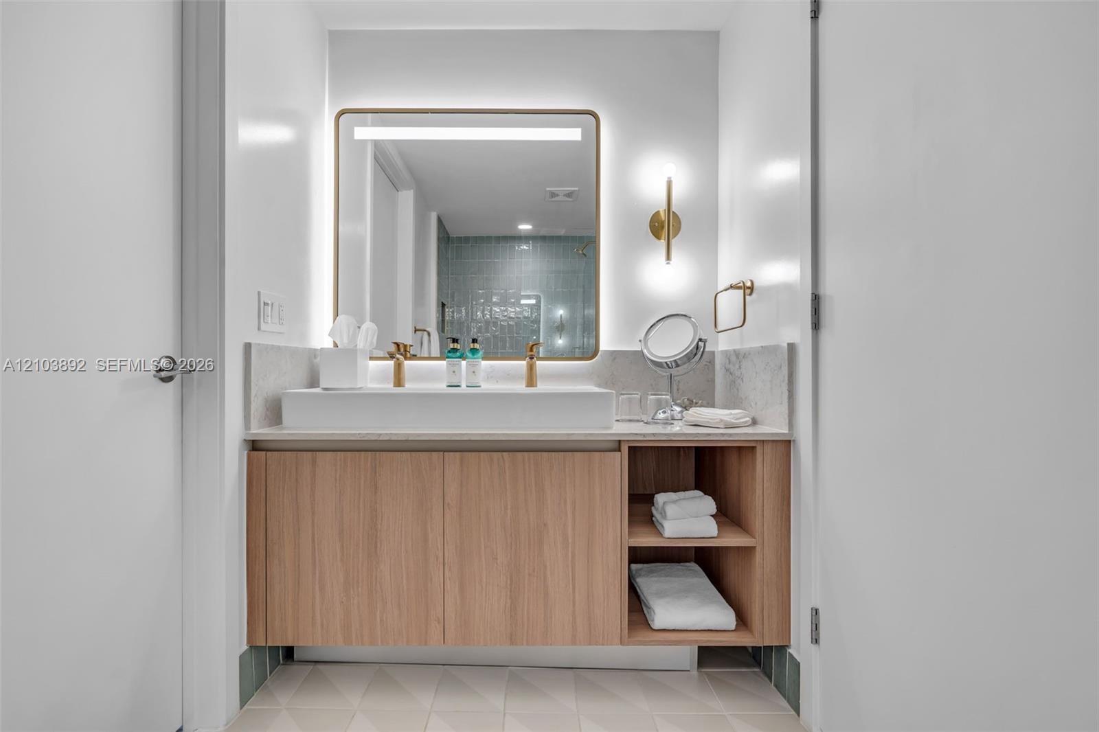 Floating vanity featuring wood-finish cabinetry with open shelving, rectangular vessel sink, stone countertop, integrated LED-backlit mirror, and metallic wall-mounted light fixtures