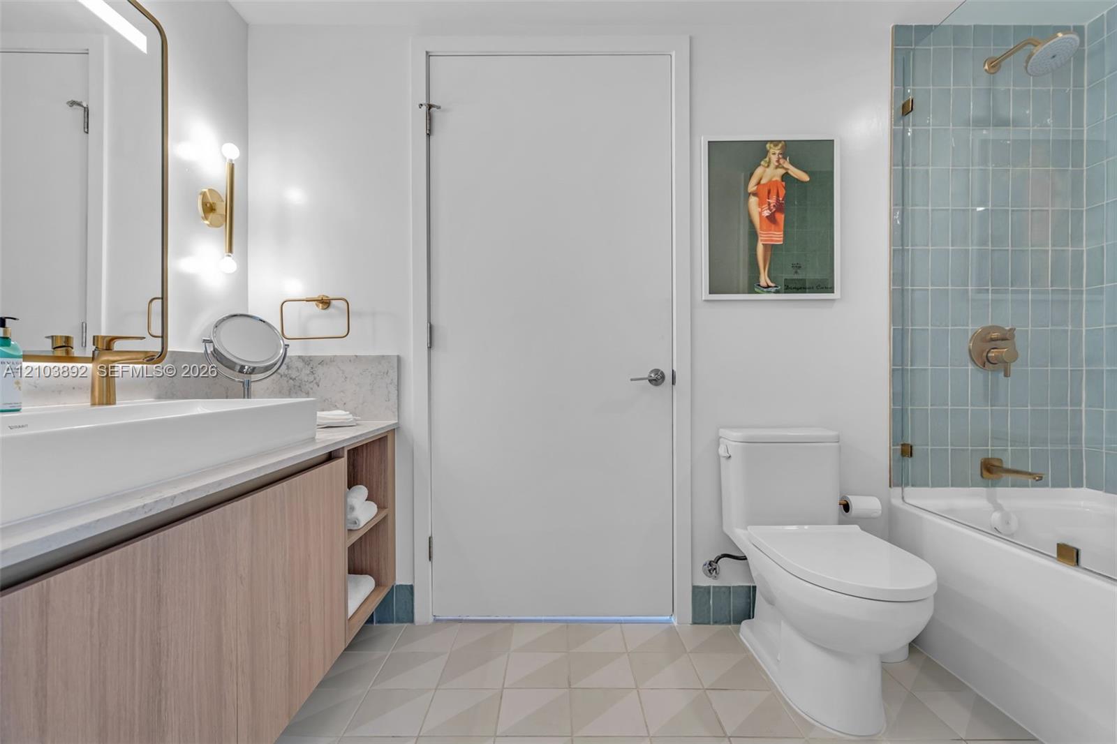 Bathroom featuring a light wood-finish vanity with an integrated countertop, wall-mounted brass hardware, a glass-enclosed shower with blue tile surround, and geometric patterned floor tile