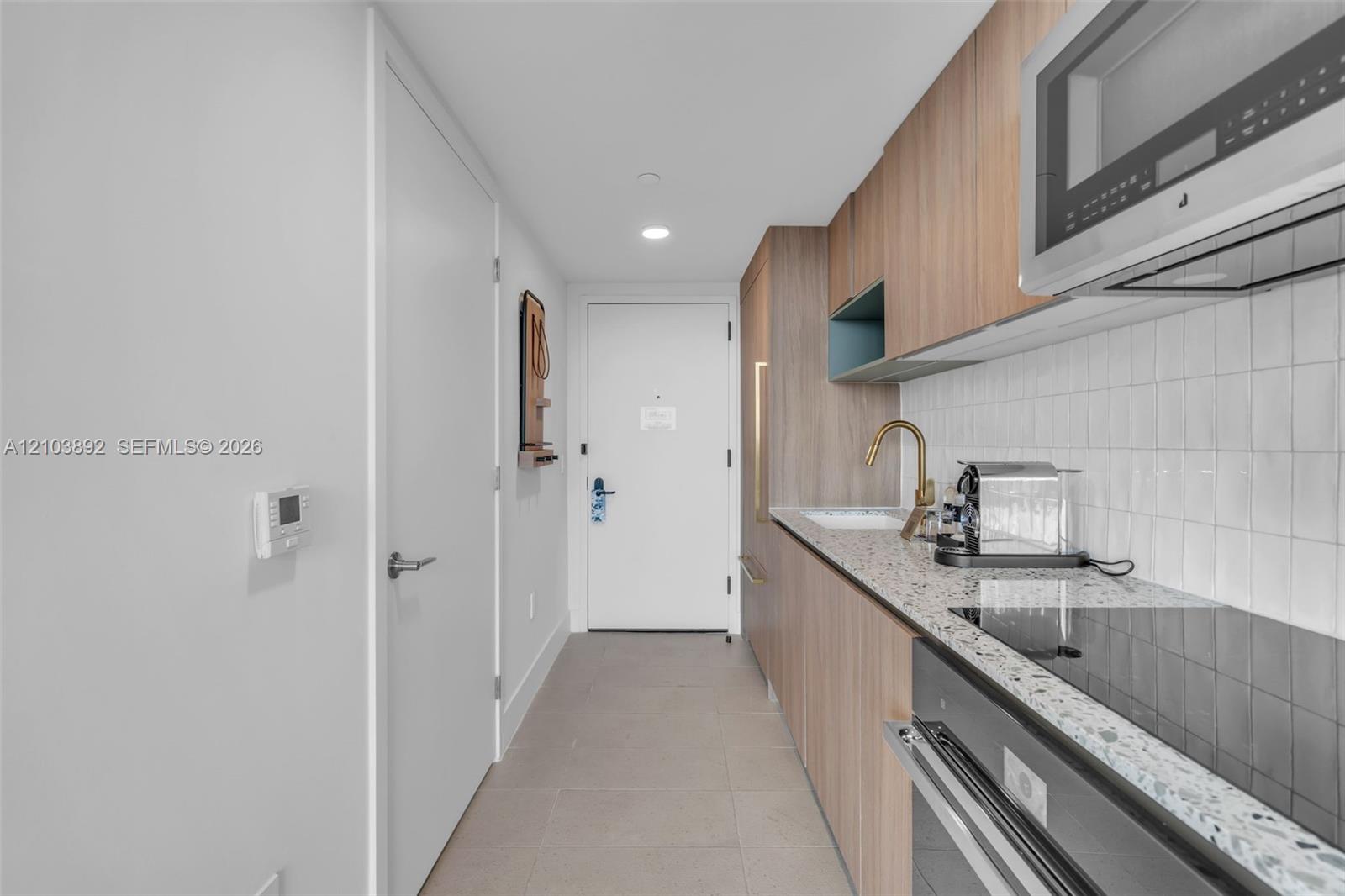 Kitchenette featuring flat-panel wood-finish cabinetry, speckled stone countertop, vertically stacked tile backsplash, brass-toned faucet, and integrated stainless steel appliances