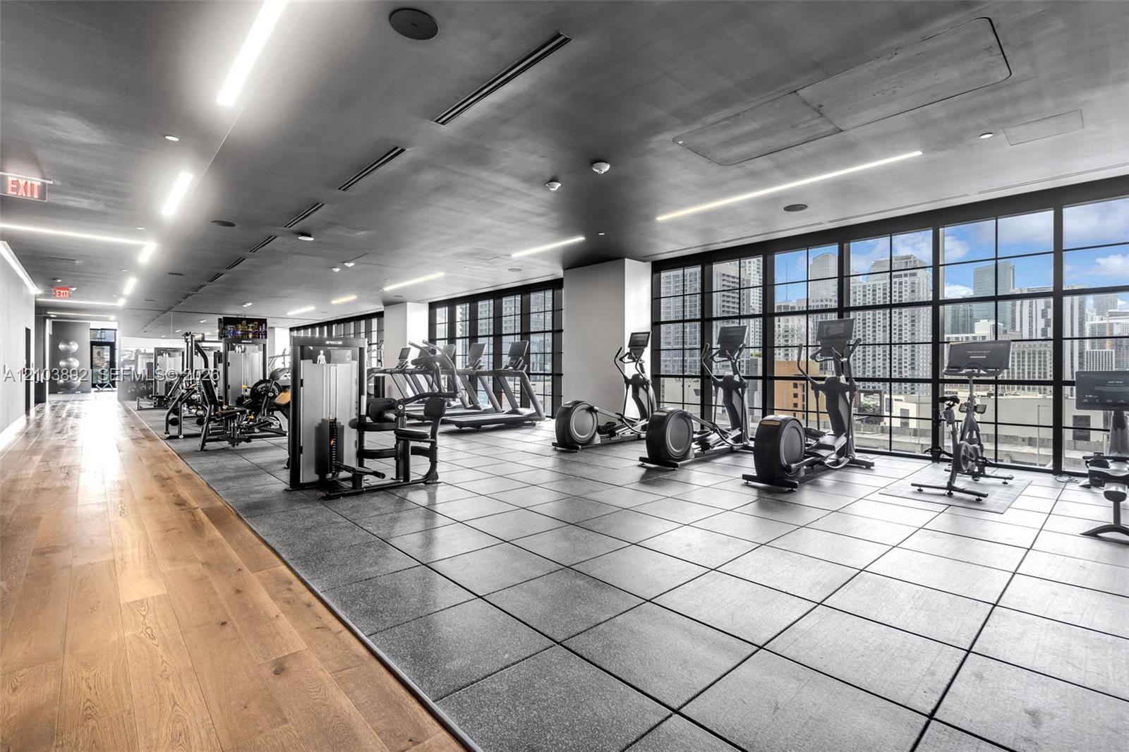 Fitness center featuring floor-to-ceiling industrial-style windows, wood-finish flooring, modular rubber tile flooring, recessed linear lighting, and integrated ceiling speakers