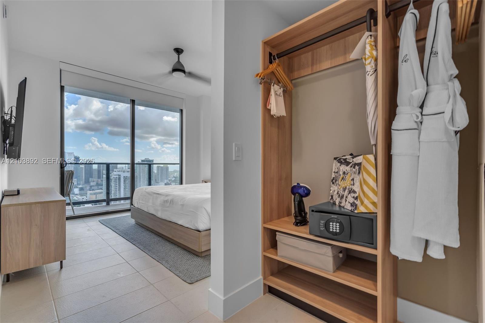 Built-in wood-finish shelving and hanging storage, floor-to-ceiling glass sliding doors, private balcony, ceiling fan, and large-format tile flooring