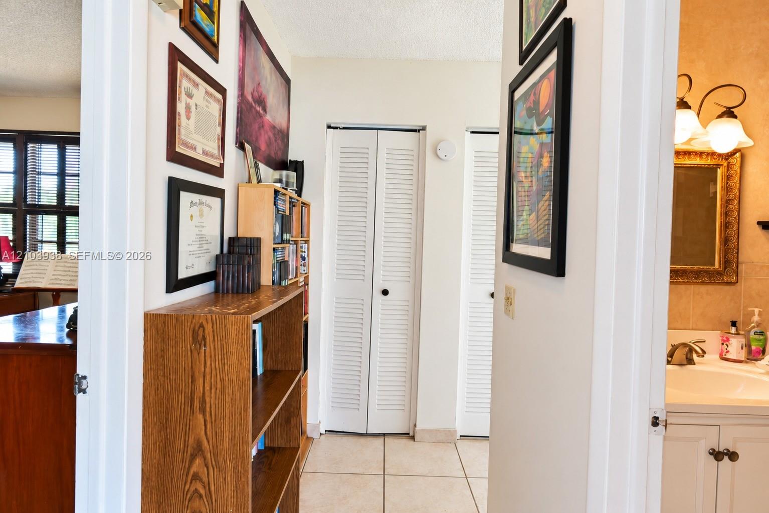 Hallway featuring light-toned tile flooring, dual white louvered-panel closet doors, and a vanity area equipped with a wall-mounted light fixture and stone-surface countertop