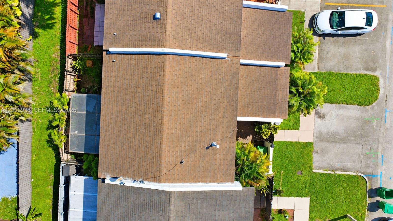 Aerial perspective of multi-pitch shingle roof featuring an enclosed patio structure, mature perimeter landscaping, concrete hardscaping, and dedicated parking