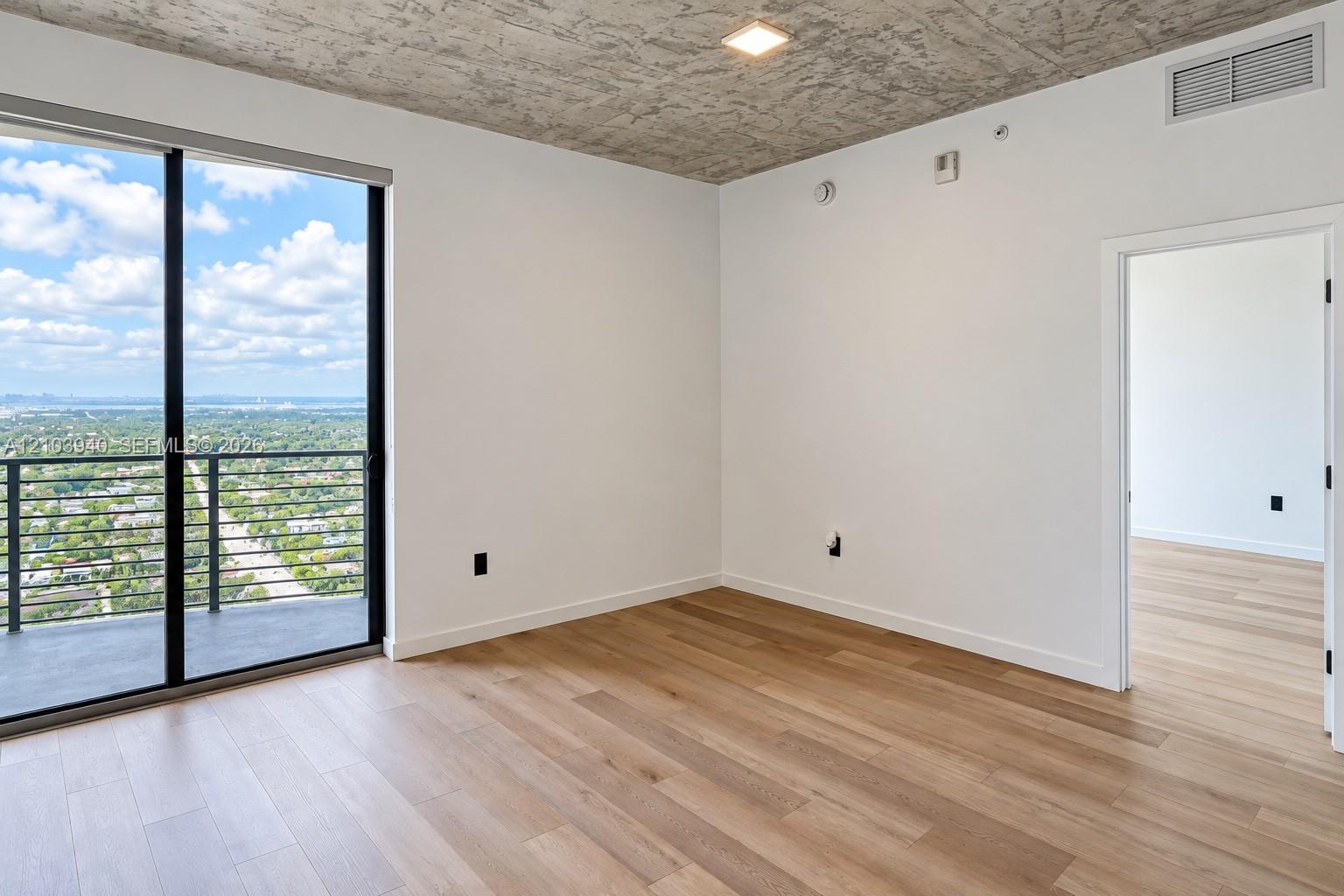 Interior space featuring raw concrete ceiling, wood-finish flooring, floor-to-ceiling glass sliding door, and private balcony with metal railing