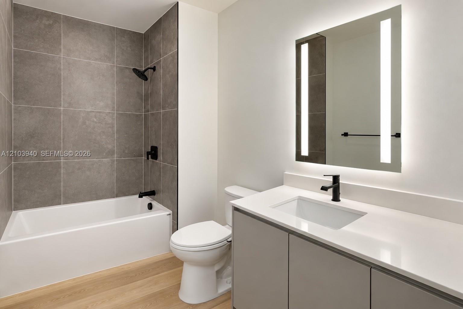Bathroom featuring a vanity with a white countertop and integrated sink, a rectangular LED-backlit mirror, wood-finish flooring, and a tub-shower combination with floor-to-ceiling grey tile surround and matte black fixtures