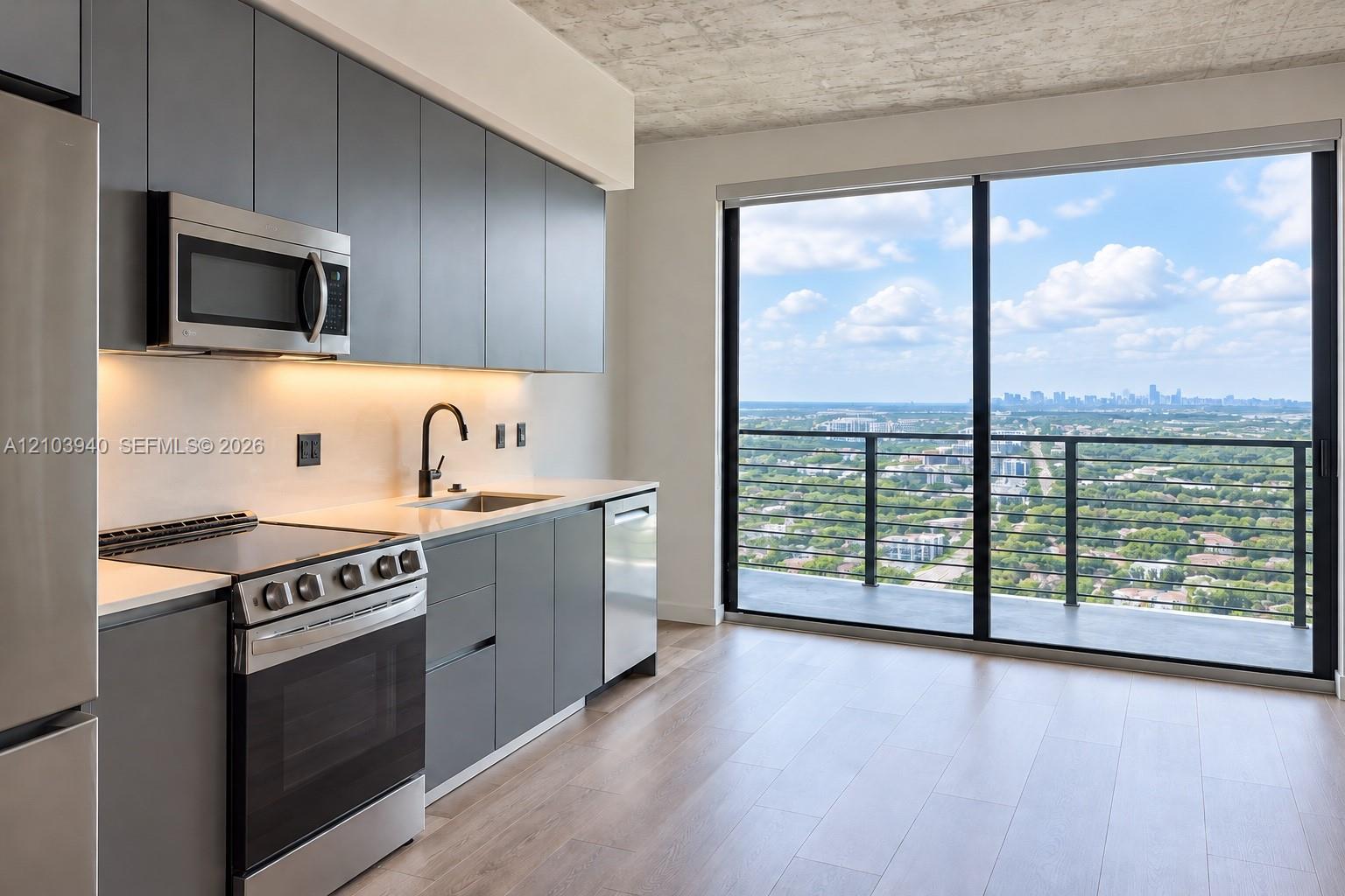 Modern kitchen featuring matte grey flat-panel cabinetry, light-toned countertops, stainless steel appliances, wood-finish flooring, and floor-to-ceiling glass doors opening to a private balcony