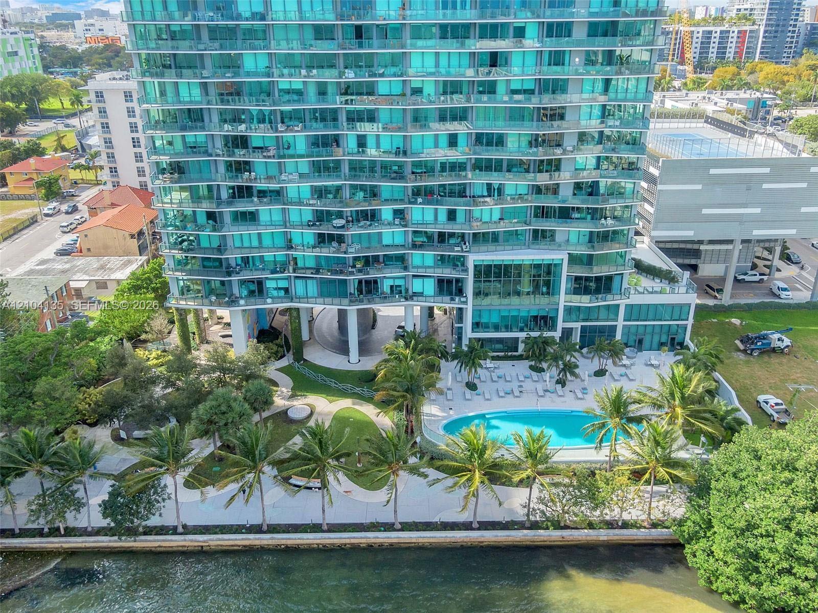High-rise glass facade building featuring wrap-around balconies, an expansive waterfront swimming pool, manicured lawn with mature palm trees, and a concrete-paved outdoor terrace