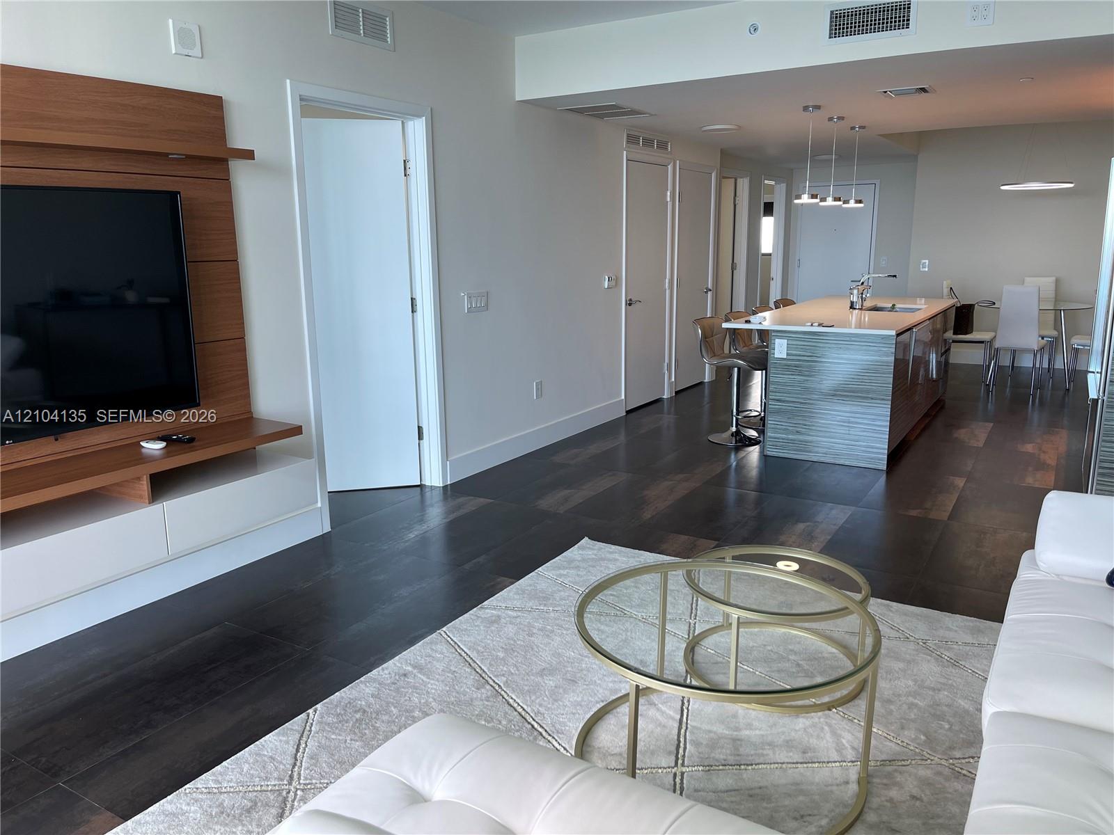 Open floor plan featuring wood-finish flooring, built-in wood-panel media console, kitchen island with stone countertop, and pendant lighting