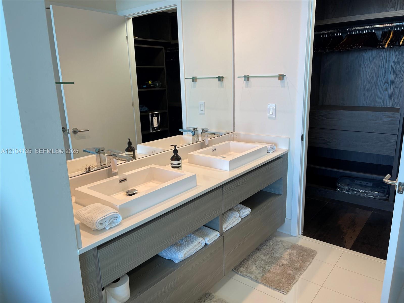 Floating dual vanity with rectangular vessel sinks, quartz countertops, and built-in shelving, complemented by a large wall-mounted mirror and walk-in closet with custom shelving