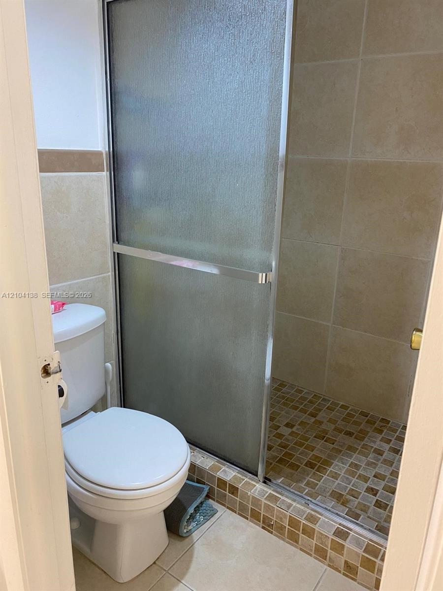 Walk-in shower with a glass sliding enclosure, floor-to-ceiling neutral tile, mosaic tile shower floor, and a ceramic-finish toilet