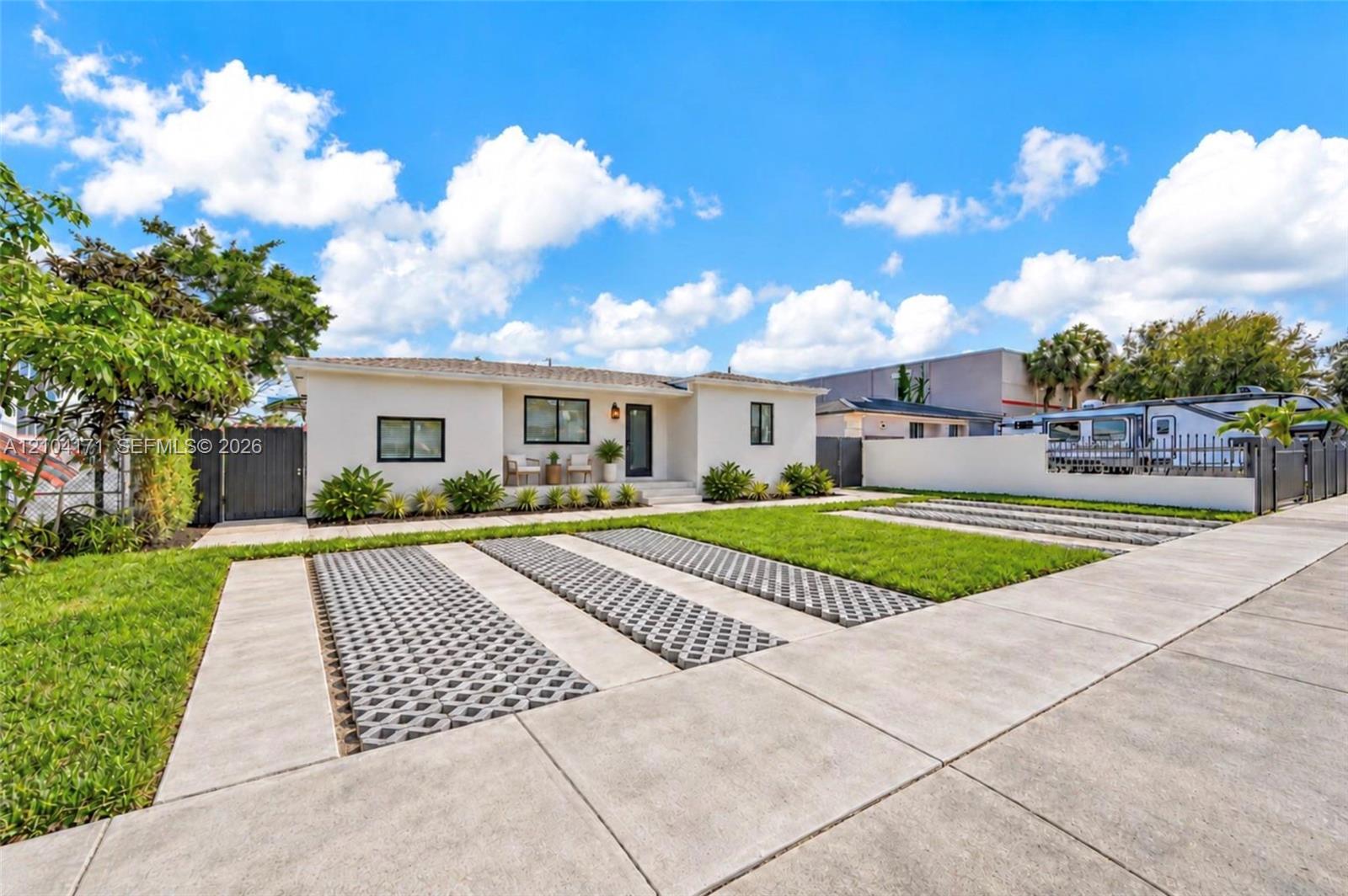 Single-story exterior featuring light-toned stucco siding, concrete ribbon driveway with integrated permeable pavers, manicured lawn, and gated perimeter fencing