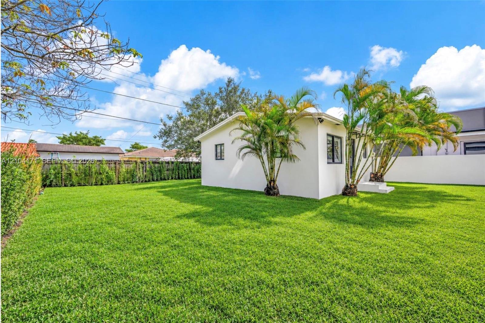 Expansive backyard featuring a manicured lawn, established palm trees, hedge fencing, and a crisp white exterior finish