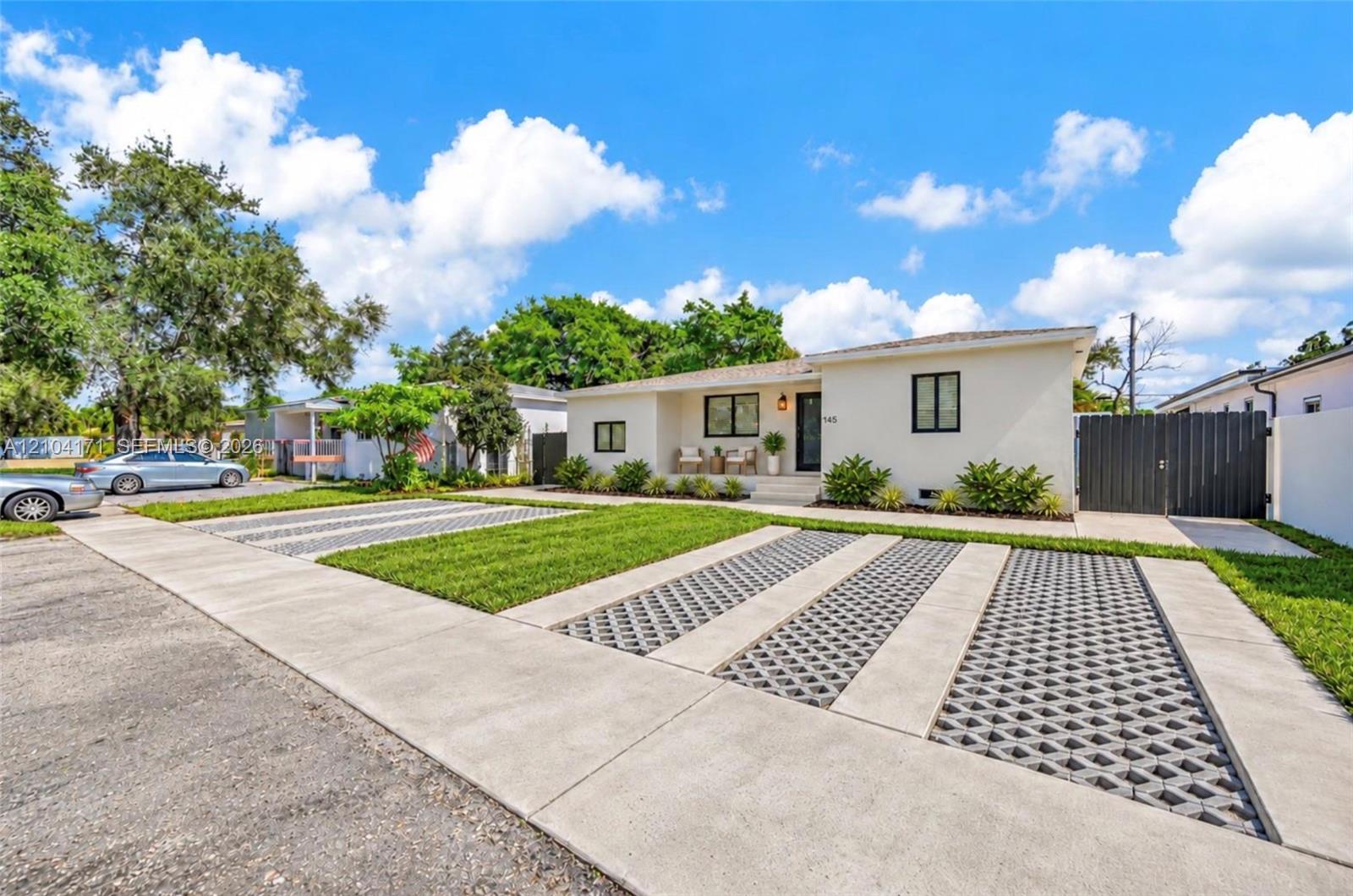Single-story residence featuring light-colored exterior siding, multi-vehicle parking area with geometric paver inserts, manicured front lawn, and dark-finished privacy fencing