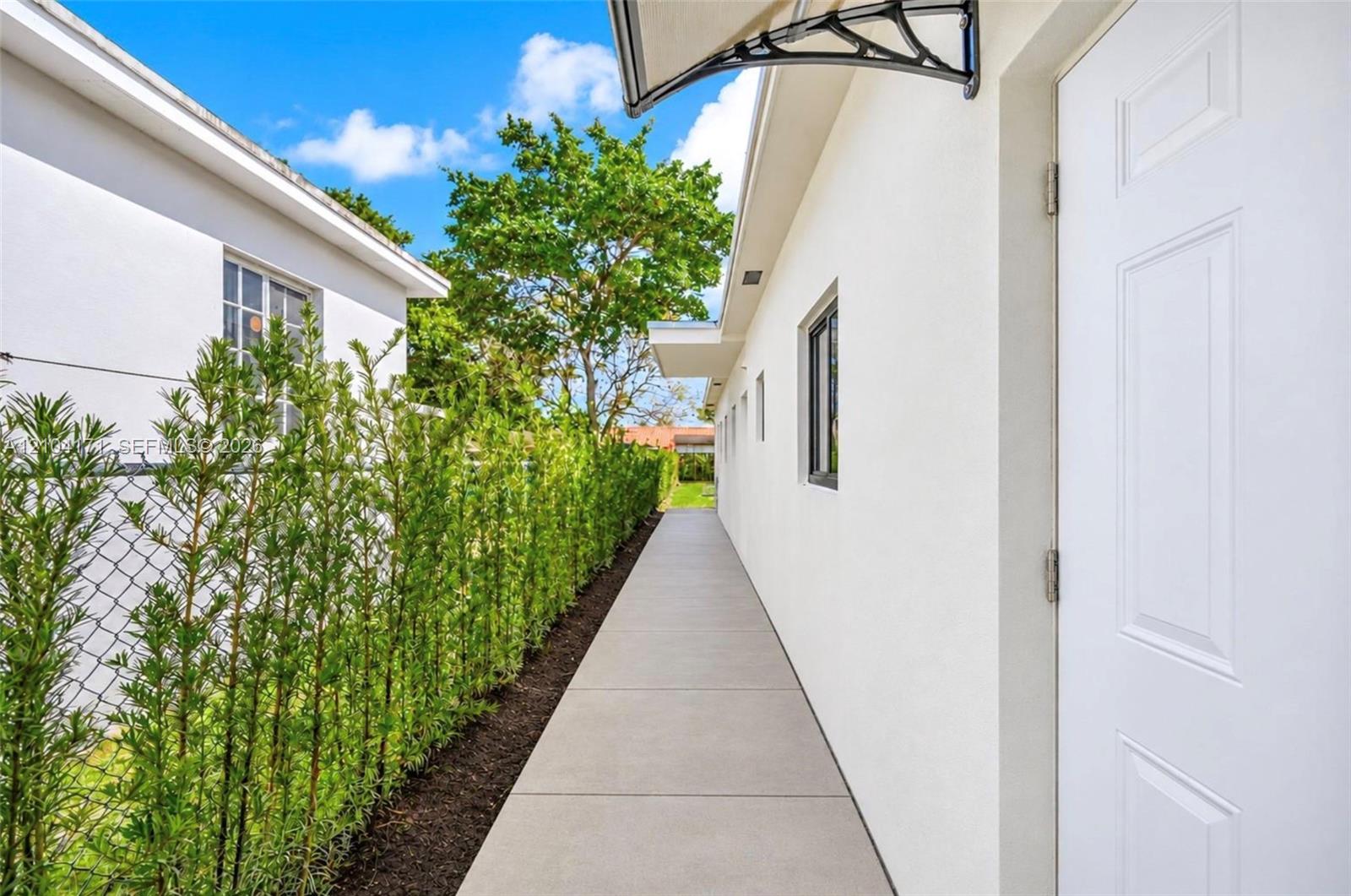 Exterior walkway featuring concrete paving, smooth stucco siding, a metal door awning, and a tall privacy hedge