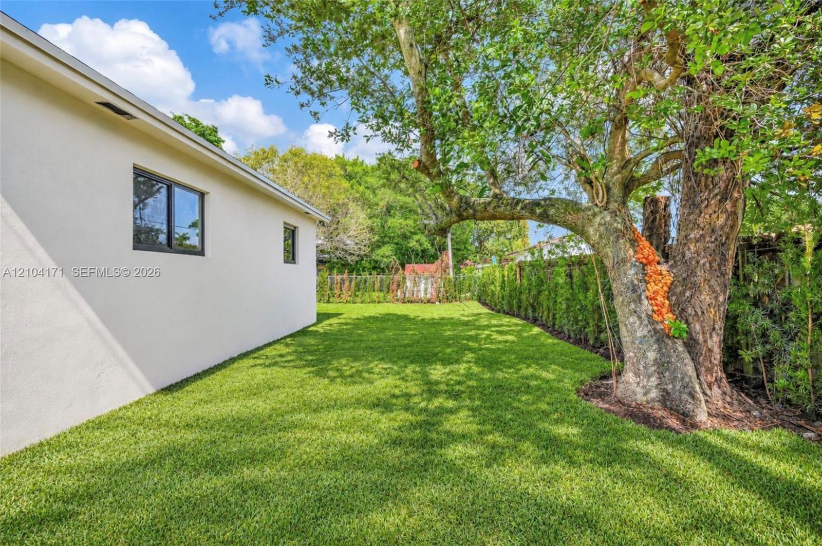Side yard featuring a smooth stucco exterior, expansive lawn, mature tree, and perimeter privacy hedges
