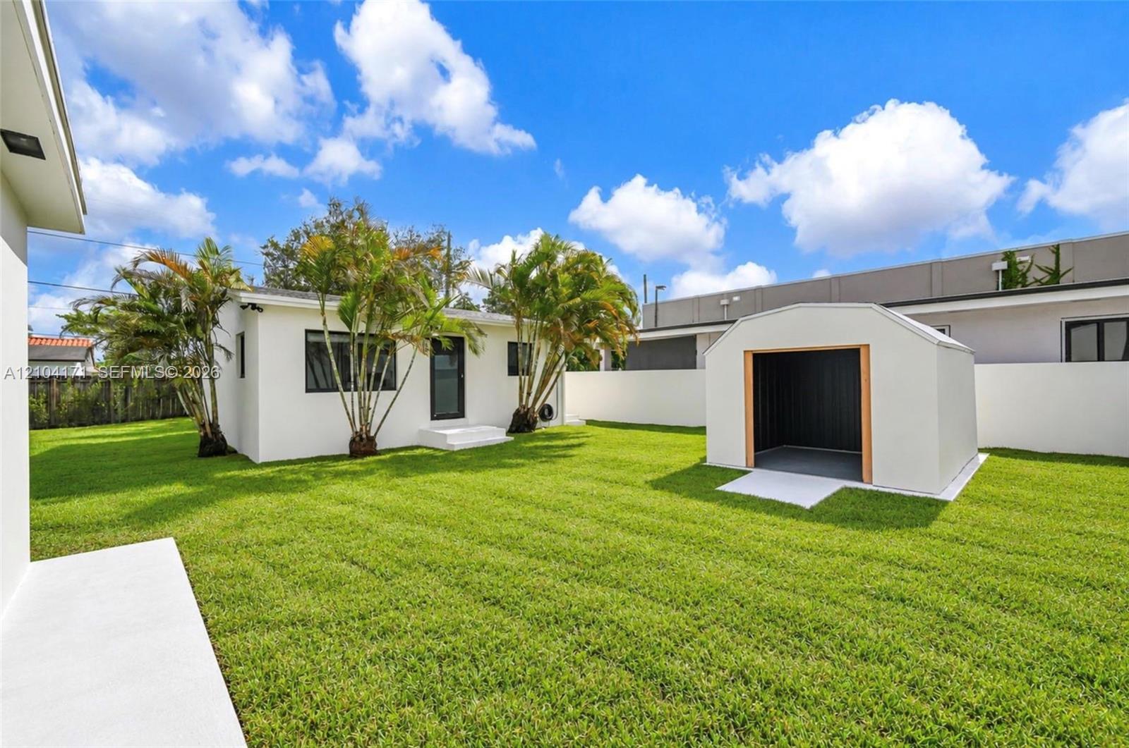 Expansive backyard featuring a manicured lawn, white perimeter wall, detached storage shed, and mature palm trees