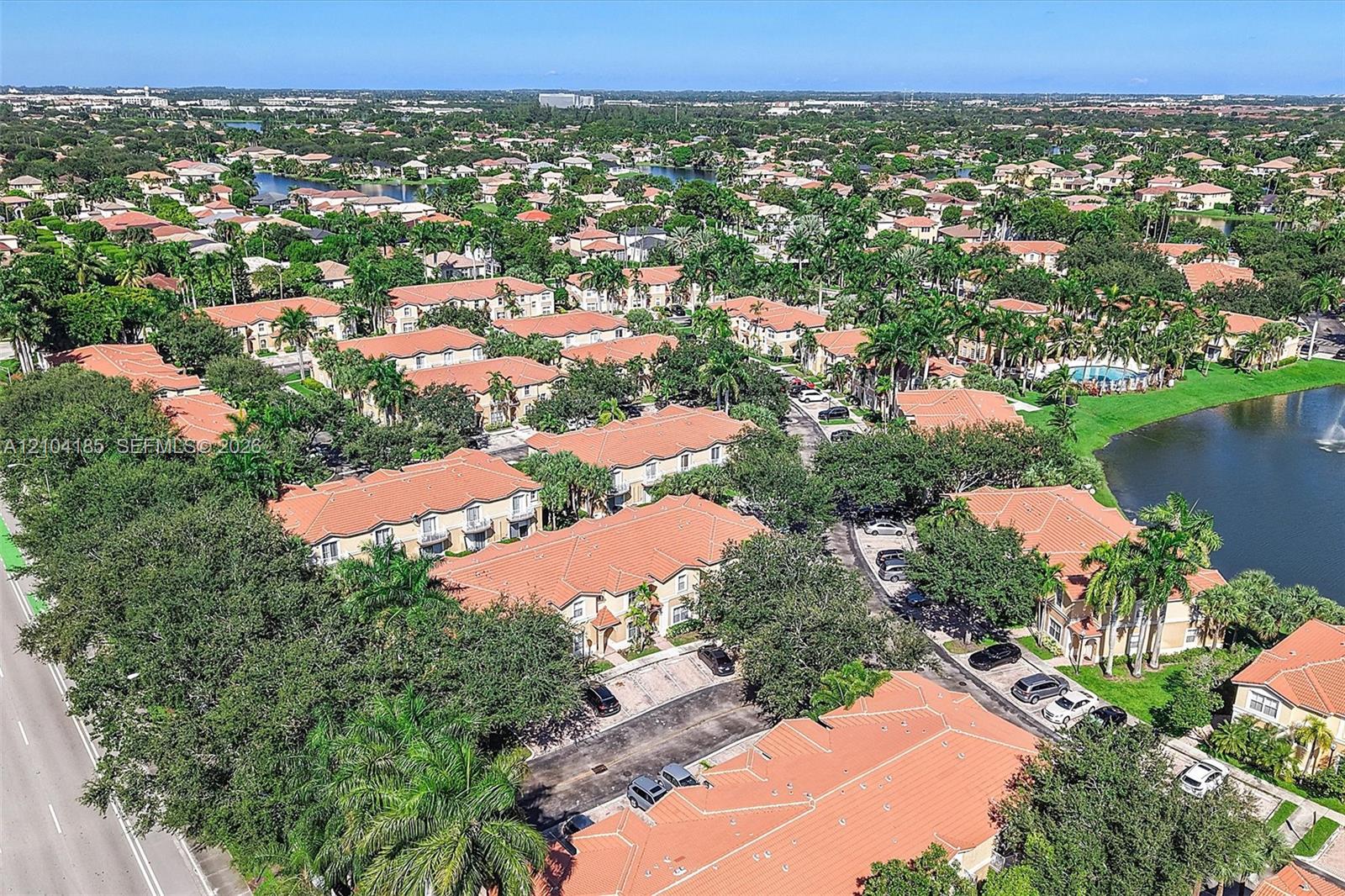 Aerial perspective of a multi-unit residential community featuring terracotta-style roofing, abundant mature landscaping, palm trees, and an on-site water feature
