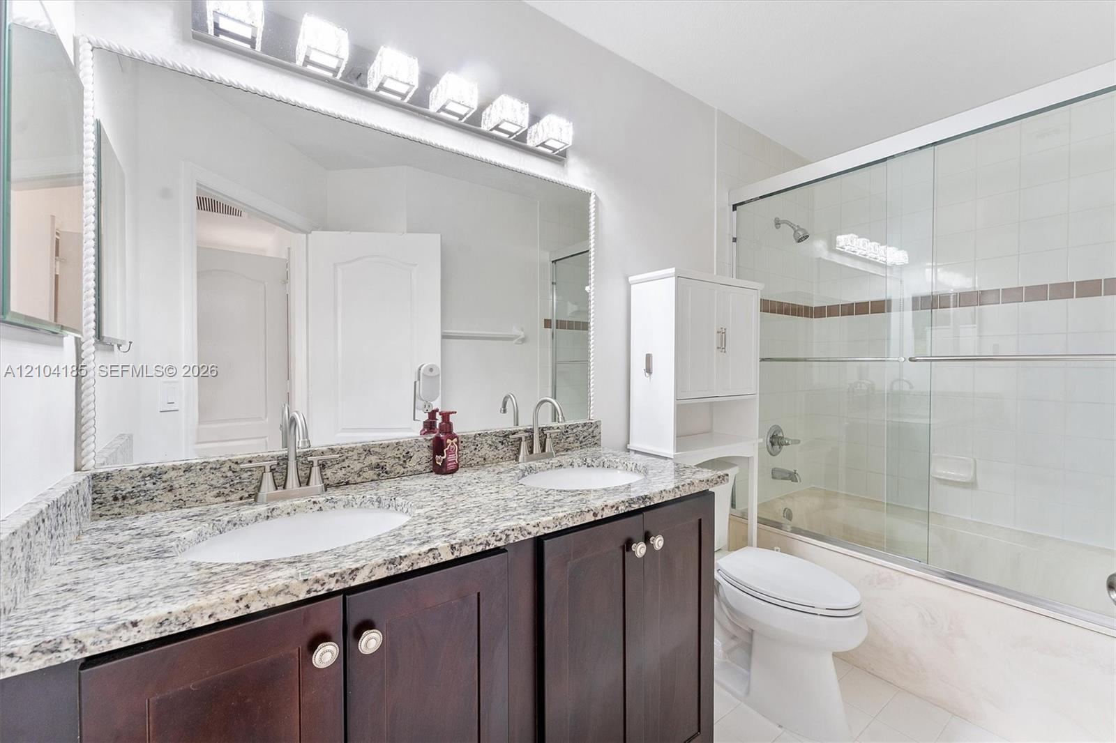 Bathroom featuring a dual-sink vanity with stone-finish countertops, dark wood-finish cabinetry, a full-width mirror, and a shower-tub combination with glass enclosure