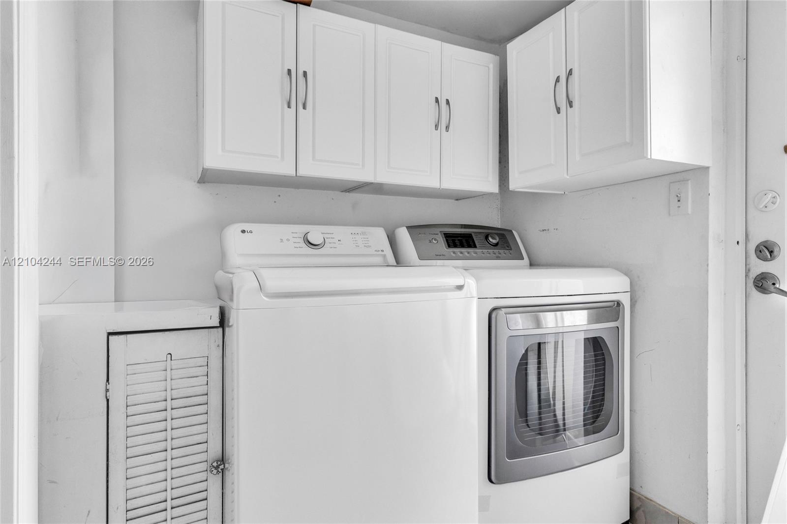 Laundry area featuring white shaker-style wall-mounted cabinetry, built-in lower storage with louvered door, and stainless steel hardware
