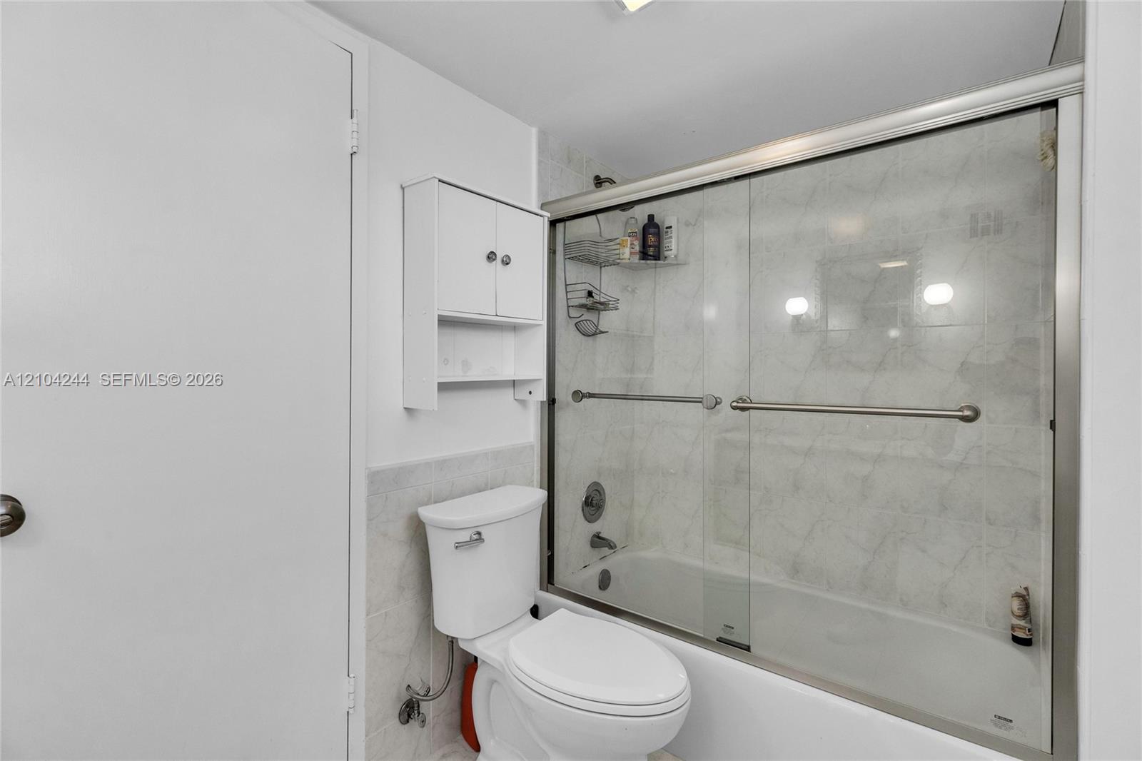 Bathroom featuring a white toilet, wall-mounted storage cabinet, and a bathtub with a glass sliding enclosure