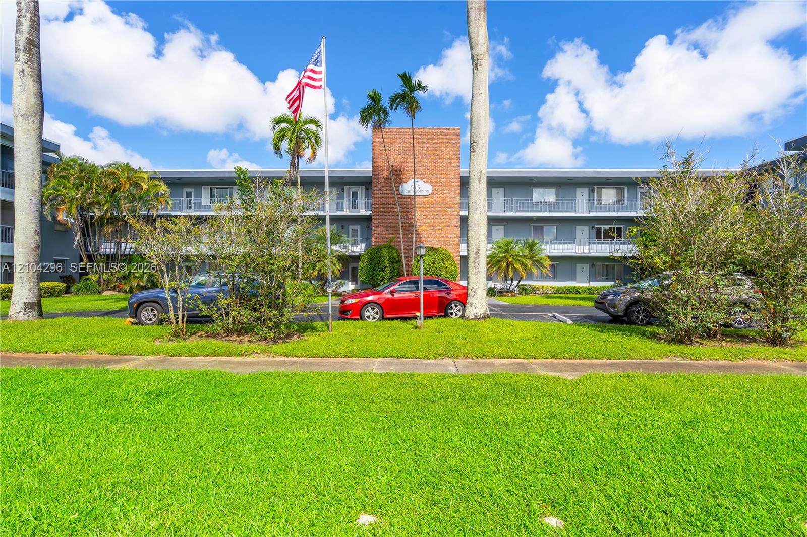 Mid-rise residential building exterior featuring a central red brick facade, grey siding, covered exterior walkways, and mature palm trees