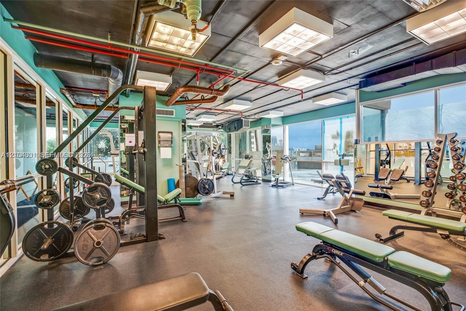 Fitness center featuring industrial-style exposed ceiling, rubberized flooring, floor-to-ceiling glass wall panels, and recessed square lighting fixtures