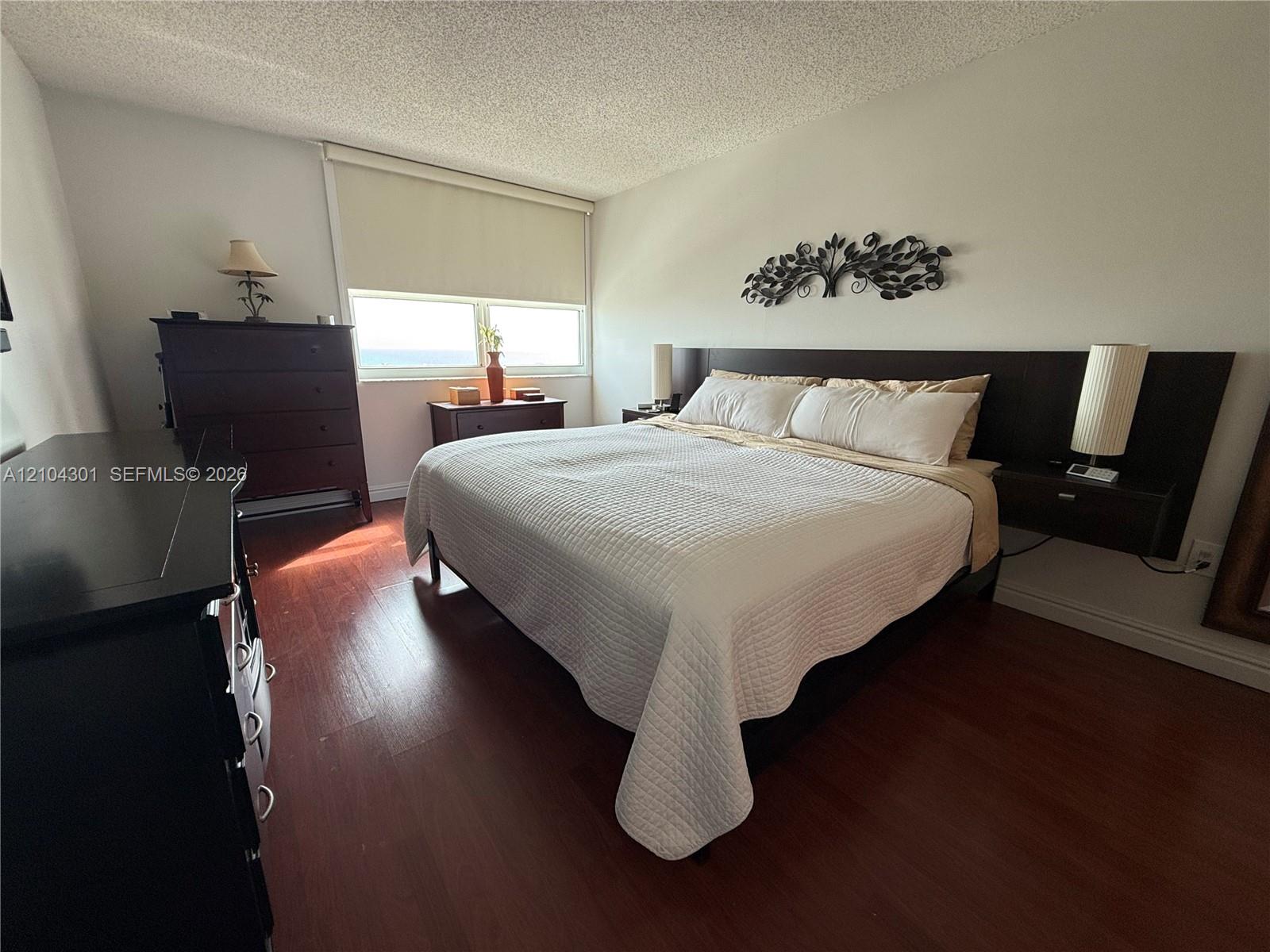Bedroom featuring wood-finish flooring, a built-in dark wood headboard with integrated floating nightstands, and large windows with roller shades Bedroom featuring wood-finish flooring, a built-in dark wood headboard with integrated floating nightstands, and large windows with roller shades