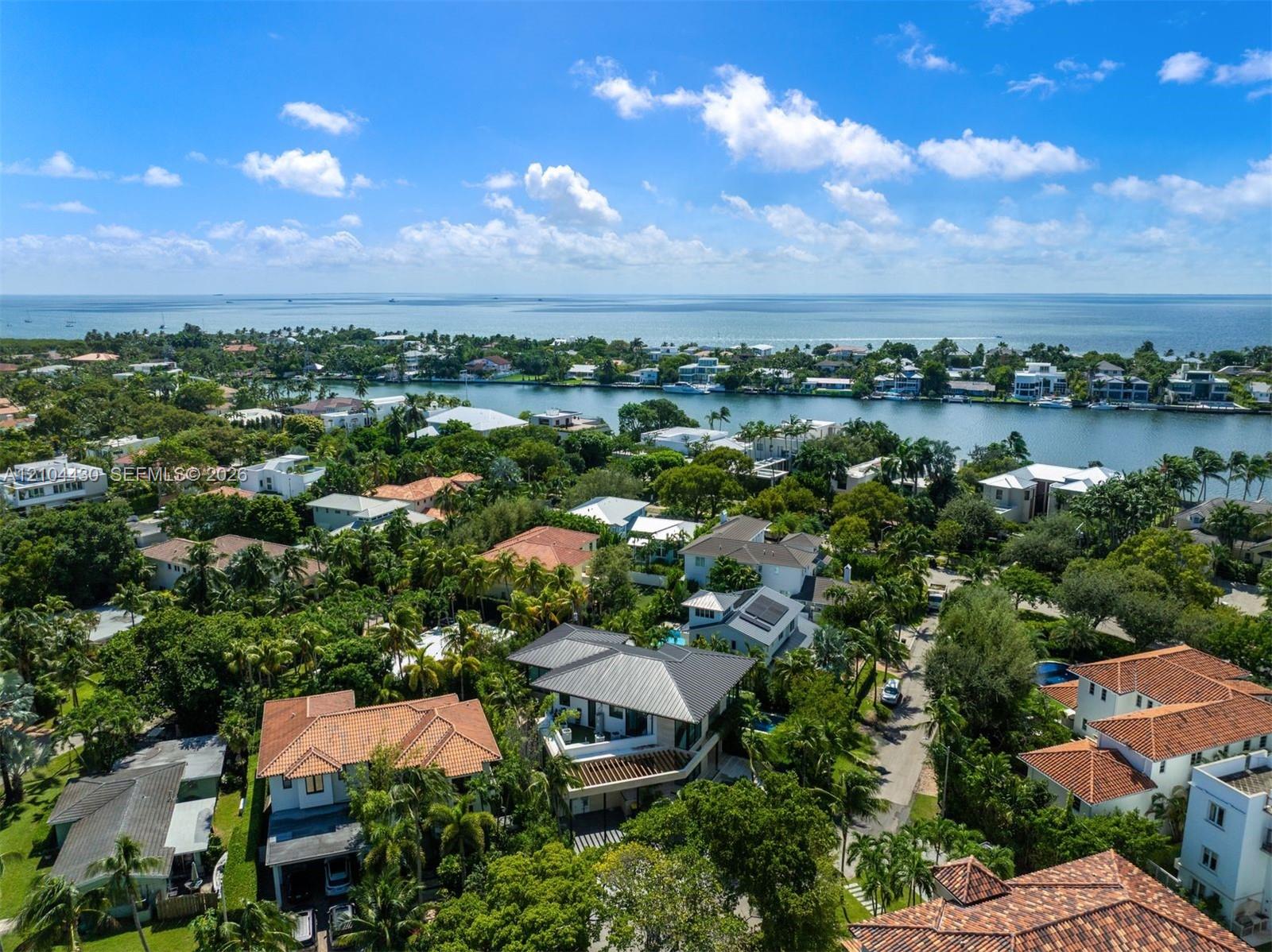 Aerial perspective of a coastal residential area featuring multi-level dwellings, terracotta tile roofing, mature canopy trees, and expansive water frontage