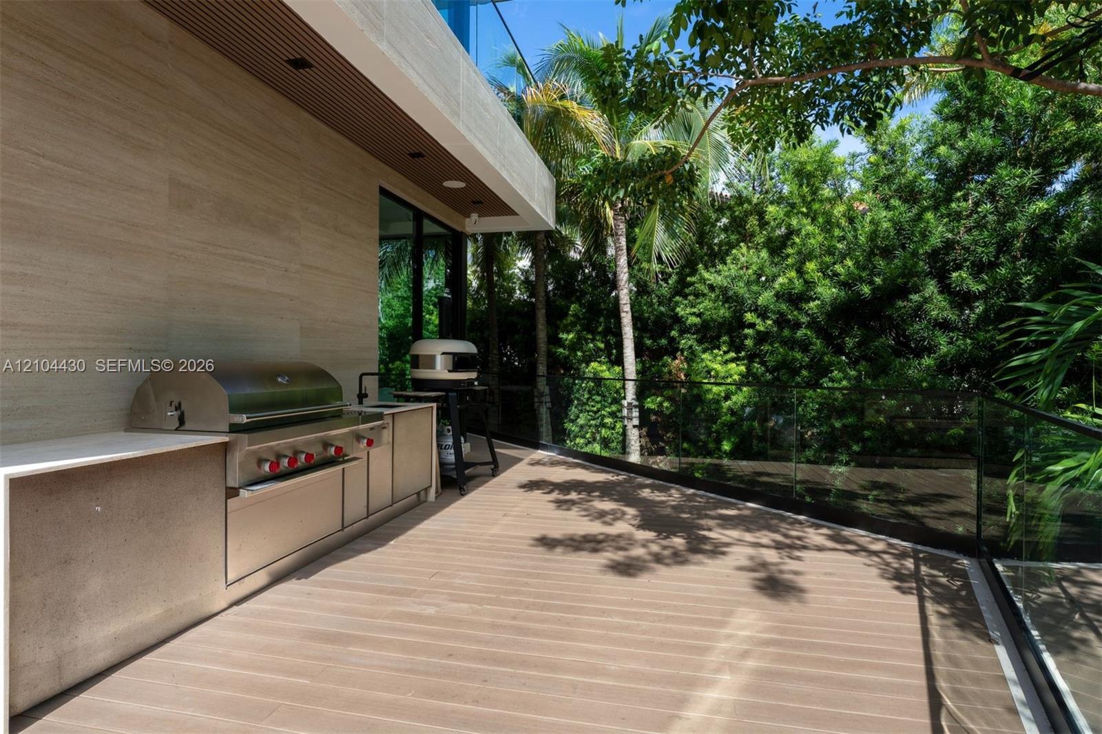 Outdoor terrace featuring wood-finish decking, glass panel railing, and a built-in outdoor kitchen with a stainless steel grill and stone-style cabinetry
