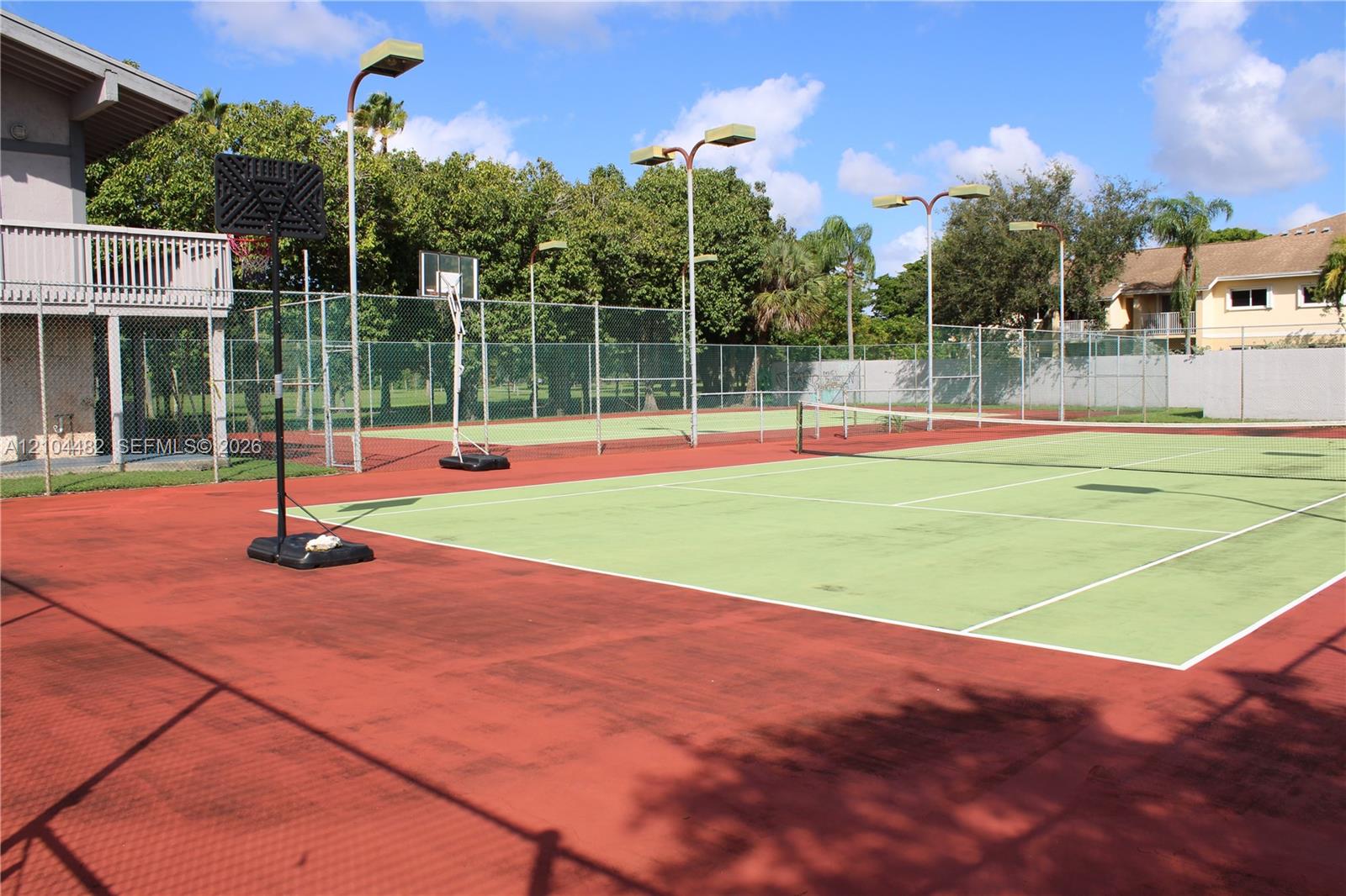 Multi-purpose sports court featuring a tennis surface, basketball hoop, perimeter chain-link fencing, and overhead court lighting