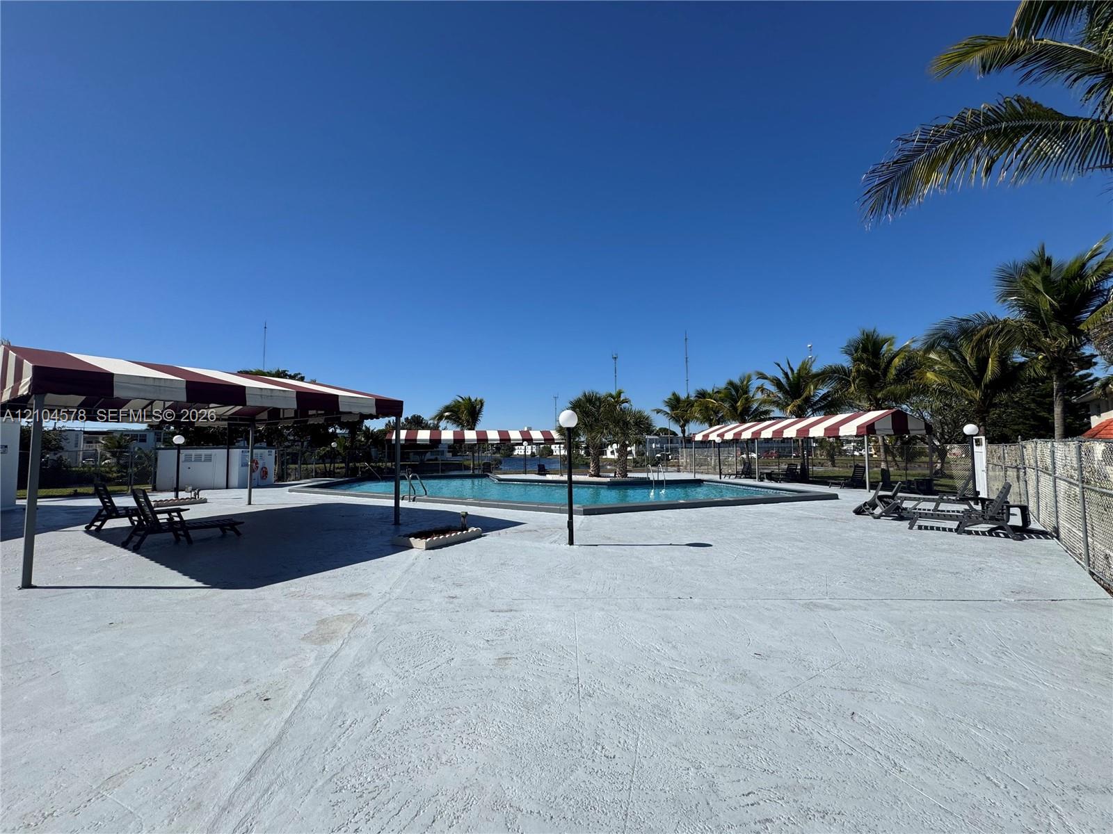 Swimming pool area featuring expansive concrete decking, multi-sectioned fabric canopies, pole-mounted globe lighting, and mature palm trees