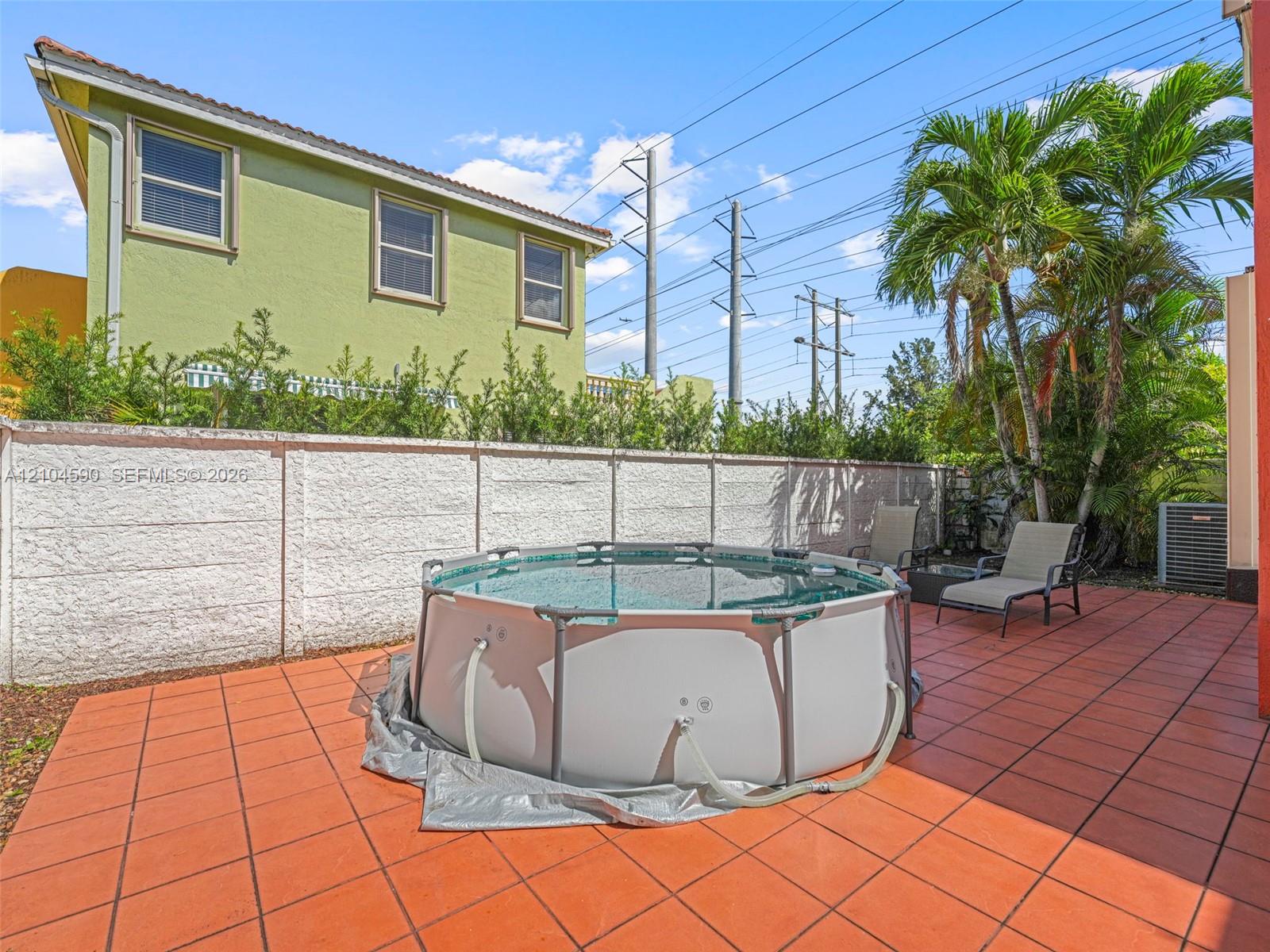 Rear exterior featuring a tiled patio, above-ground pool, privacy wall, mature palm trees, and a multi-story building exterior