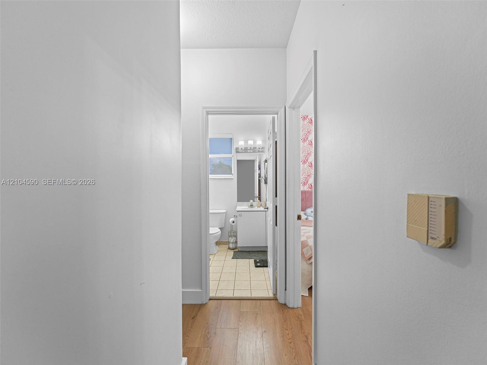 Hallway featuring wood-finish flooring, a wall-mounted climate control unit, and a doorway revealing a bathroom with tile flooring, a white vanity, and a three-light fixture