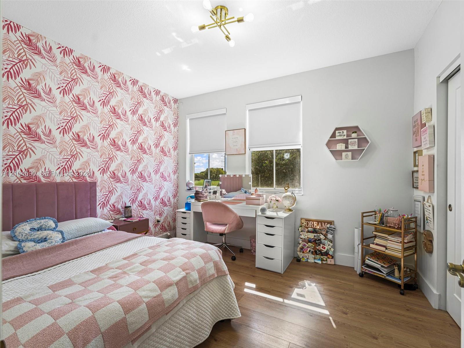 Bedroom featuring wood-finish flooring, dual windows with roller shades, a gold-tone multi-arm chandelier, and a decorative patterned accent wall Bedroom featuring wood-finish flooring, dual windows with roller shades, a gold-tone multi-arm chandelier, and a decorative patterned accent wall