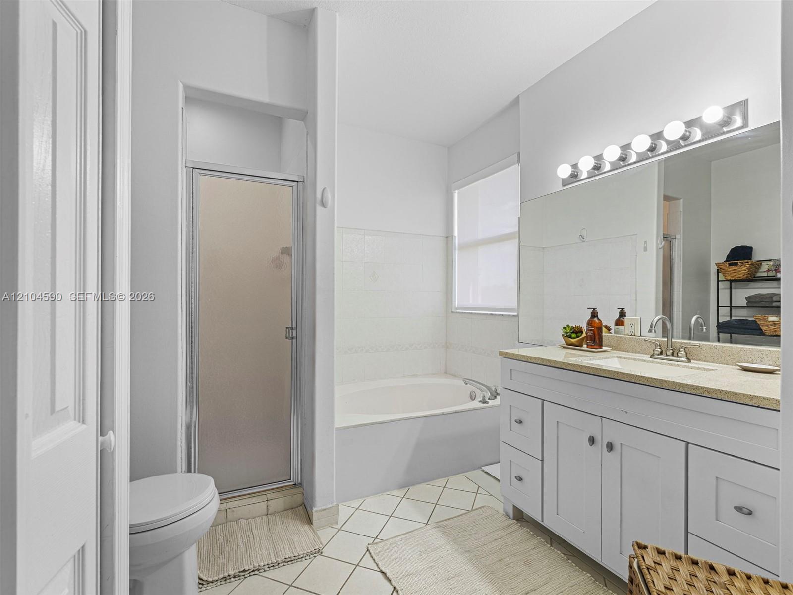 Bathroom featuring a vanity with integrated sink and stone-composite countertop, full-width wall mirror, separate shower stall with glass door, and soaking tub with tiled surround Bathroom featuring a vanity with integrated sink and stone-composite countertop, full-width wall mirror, separate shower stall with glass door, and soaking tub with tiled surround