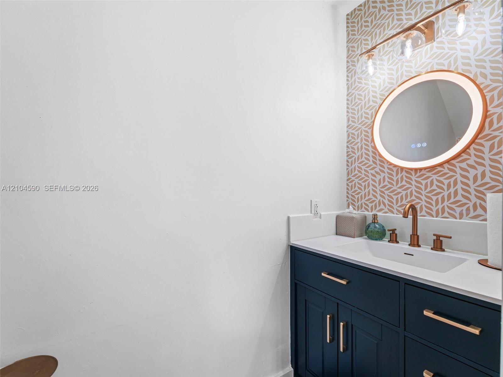 Navy blue vanity with metallic hardware, integrated white countertop, oval LED-backlit mirror, patterned accent wall, and metallic multi-light fixture