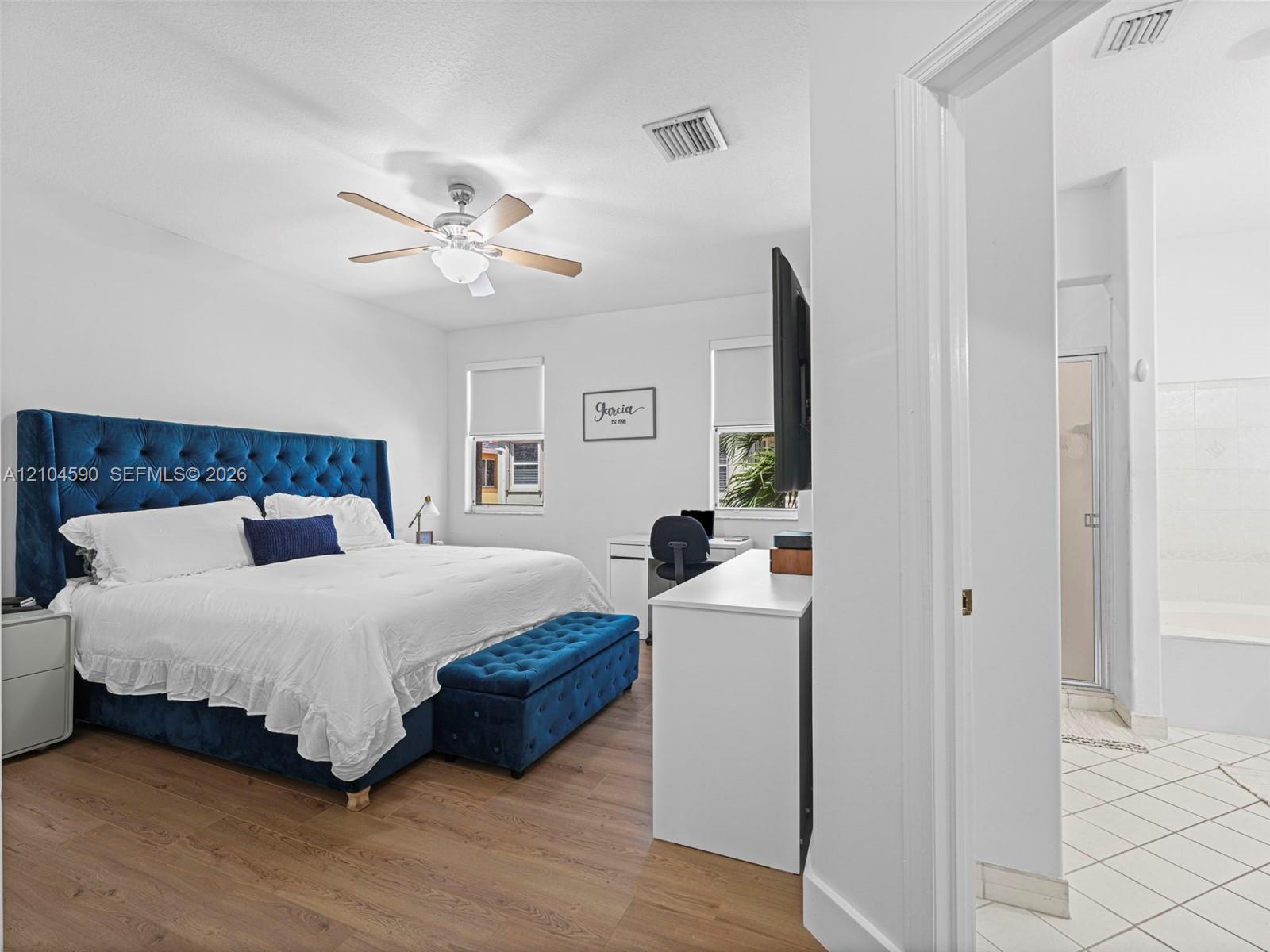 Bedroom featuring wood-finish flooring, a multi-blade ceiling fan with integrated lighting, dual windows, and an en-suite bathroom with tile flooring